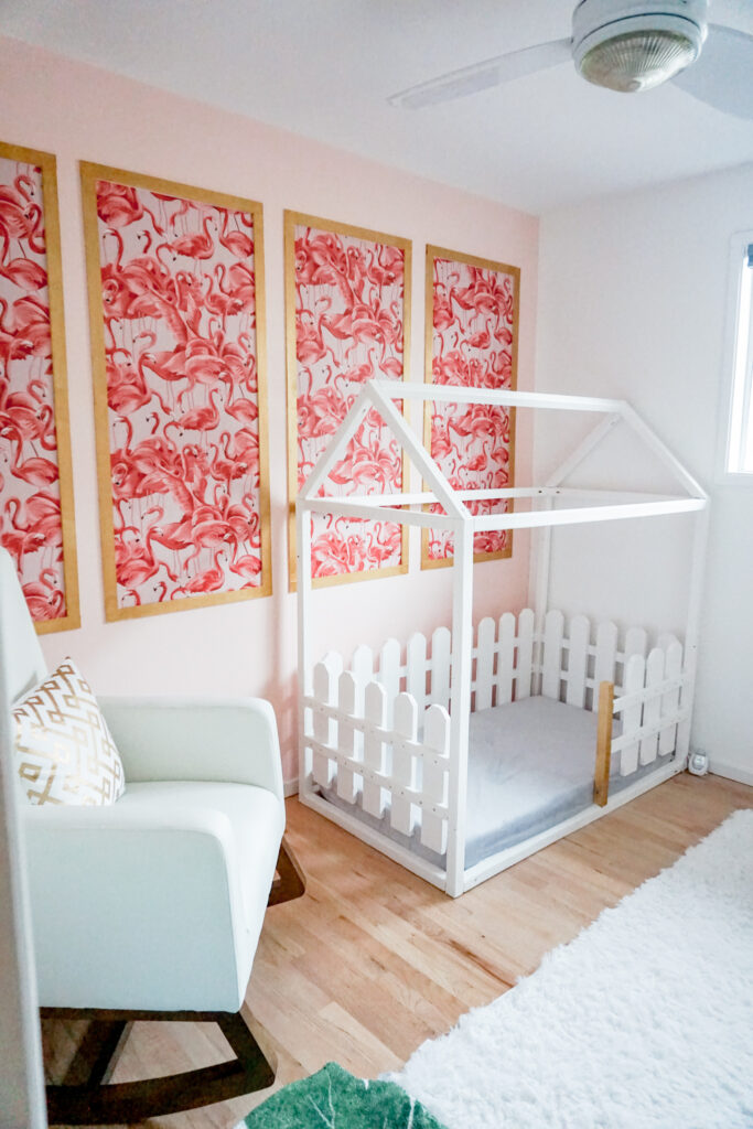 girls nursery room: flamingo wallpaper with gold frame in modern chinoiserie on a pink wall, white modern rocking chair, and white wooden house bed