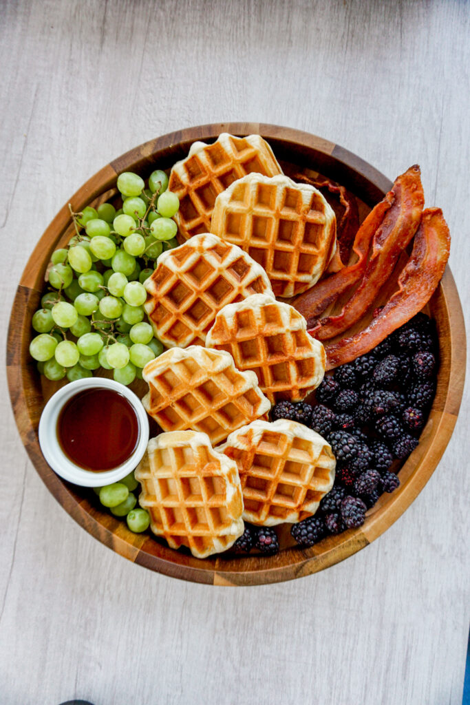 Circular charcuterie board food platter with quick Belgian waffles, bacon, syrup, grapes, and blackberries.