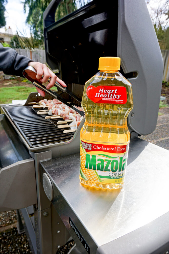 Mazola® Corn Oil in front of chicken yakitori skewers being cooked on a grill
