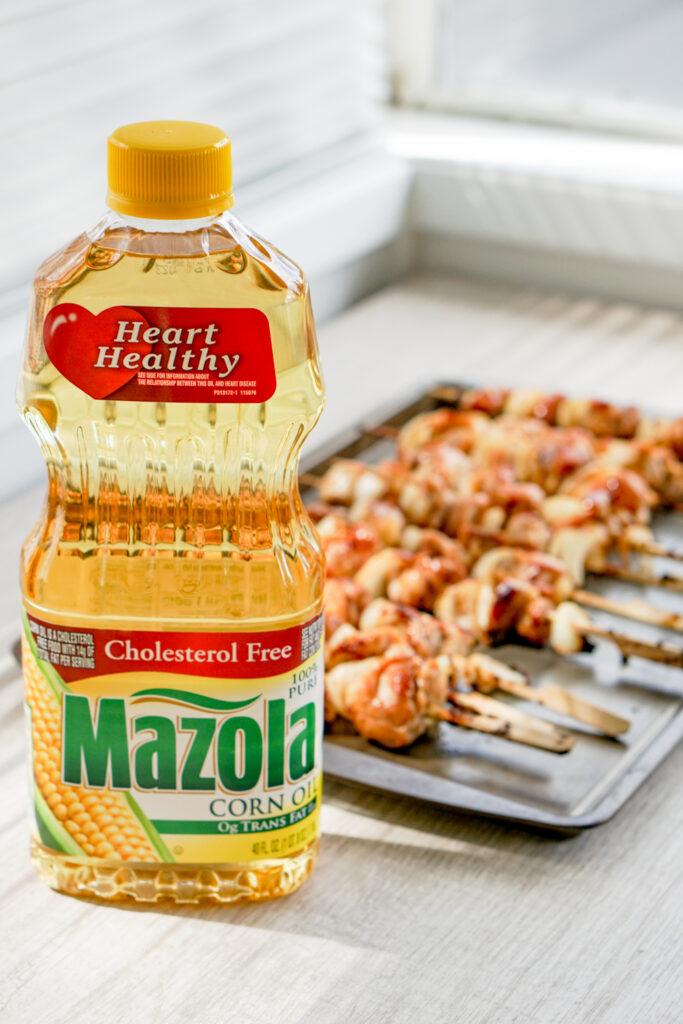 A bottle of Mazola® Corn Oil sits on a table in front of a tray of chicken yakitori skewers.